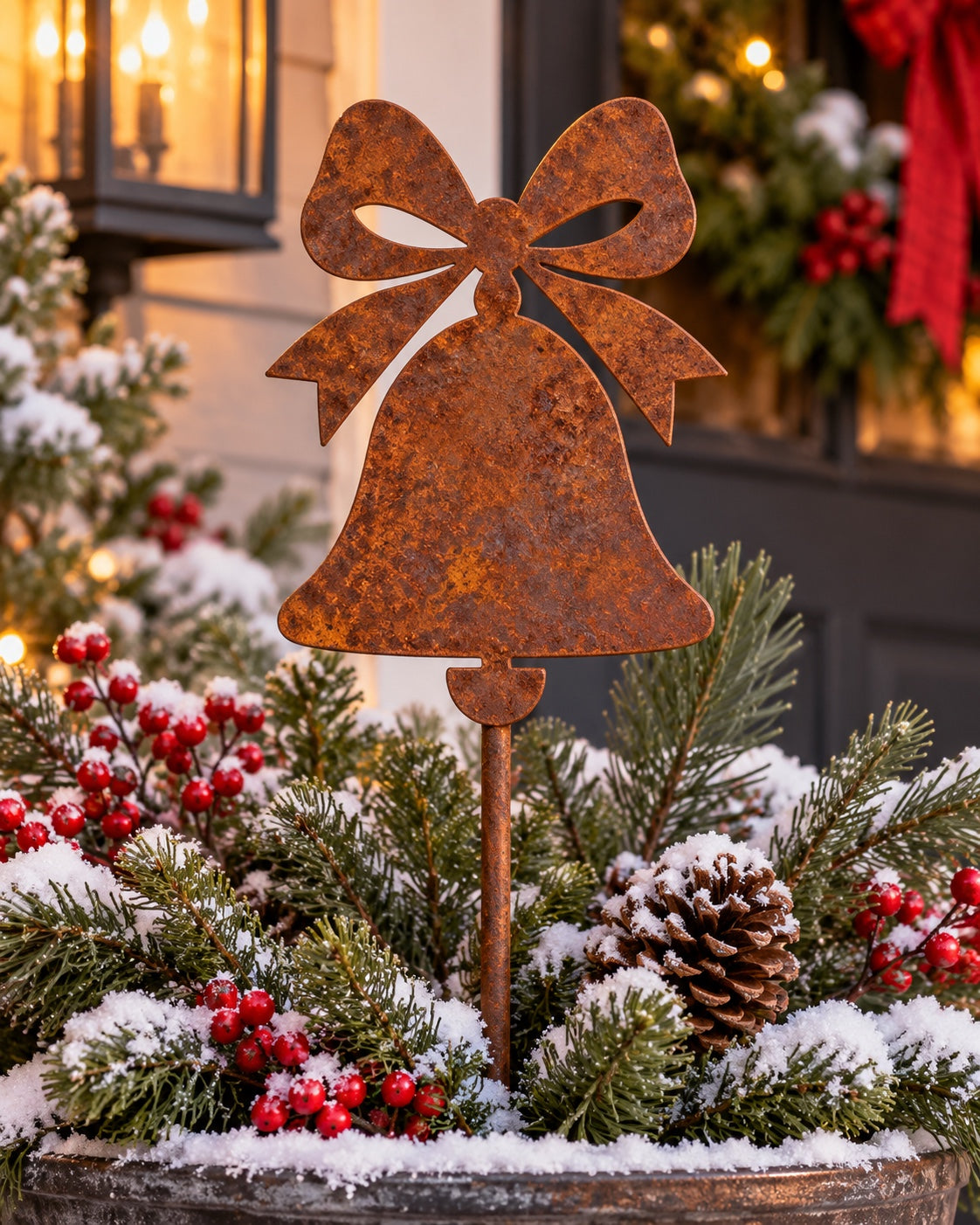 Rusted steel Christmas bell garden stake topped with a bow and open loop details, styled in a snowy evergreen planter with pinecones and red berries beside a Christmas porch.